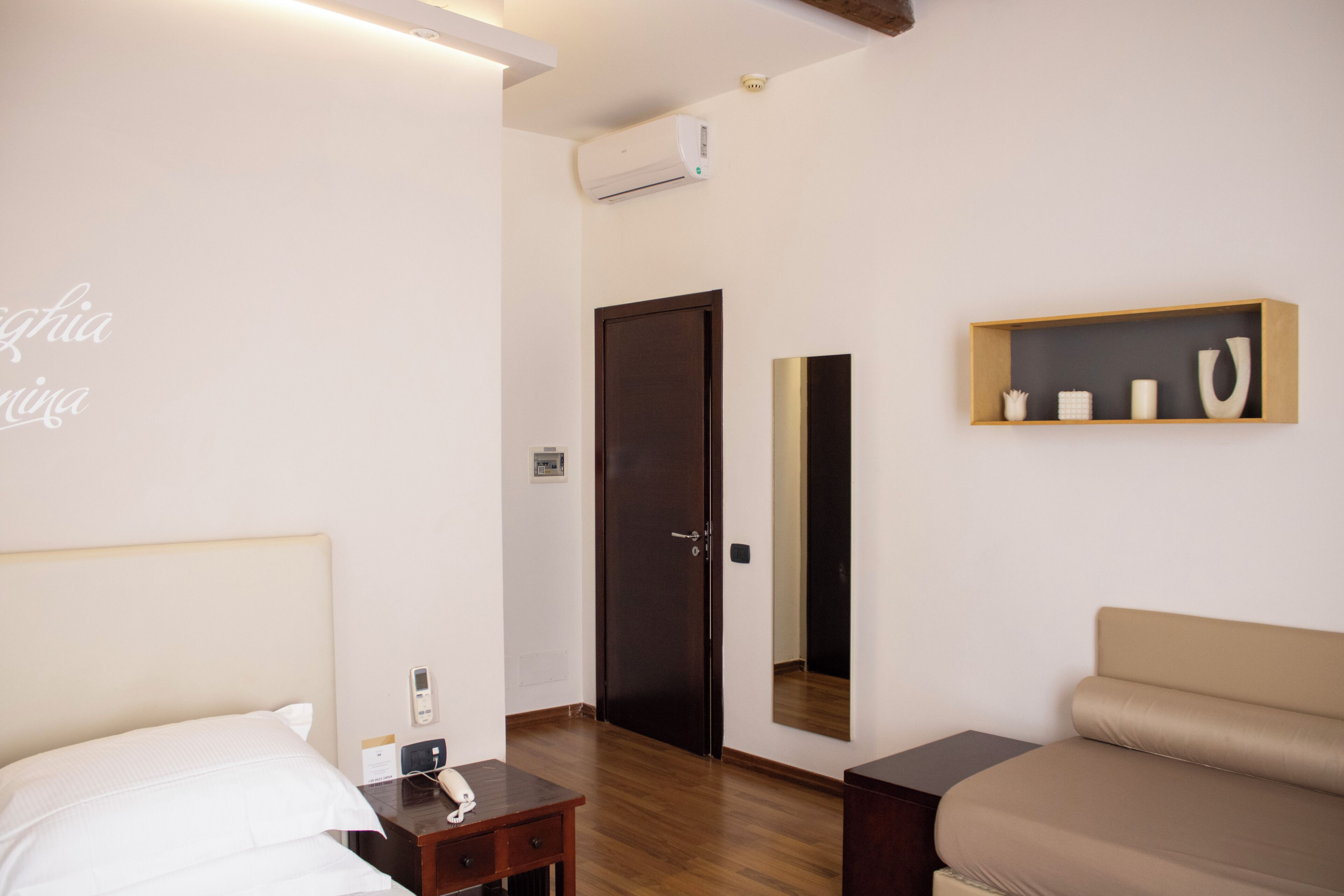 Una habitación de hotel con cama, mesita de noche, un escritorio pequeño, un sofá, un espejo y una estantería con adornos.