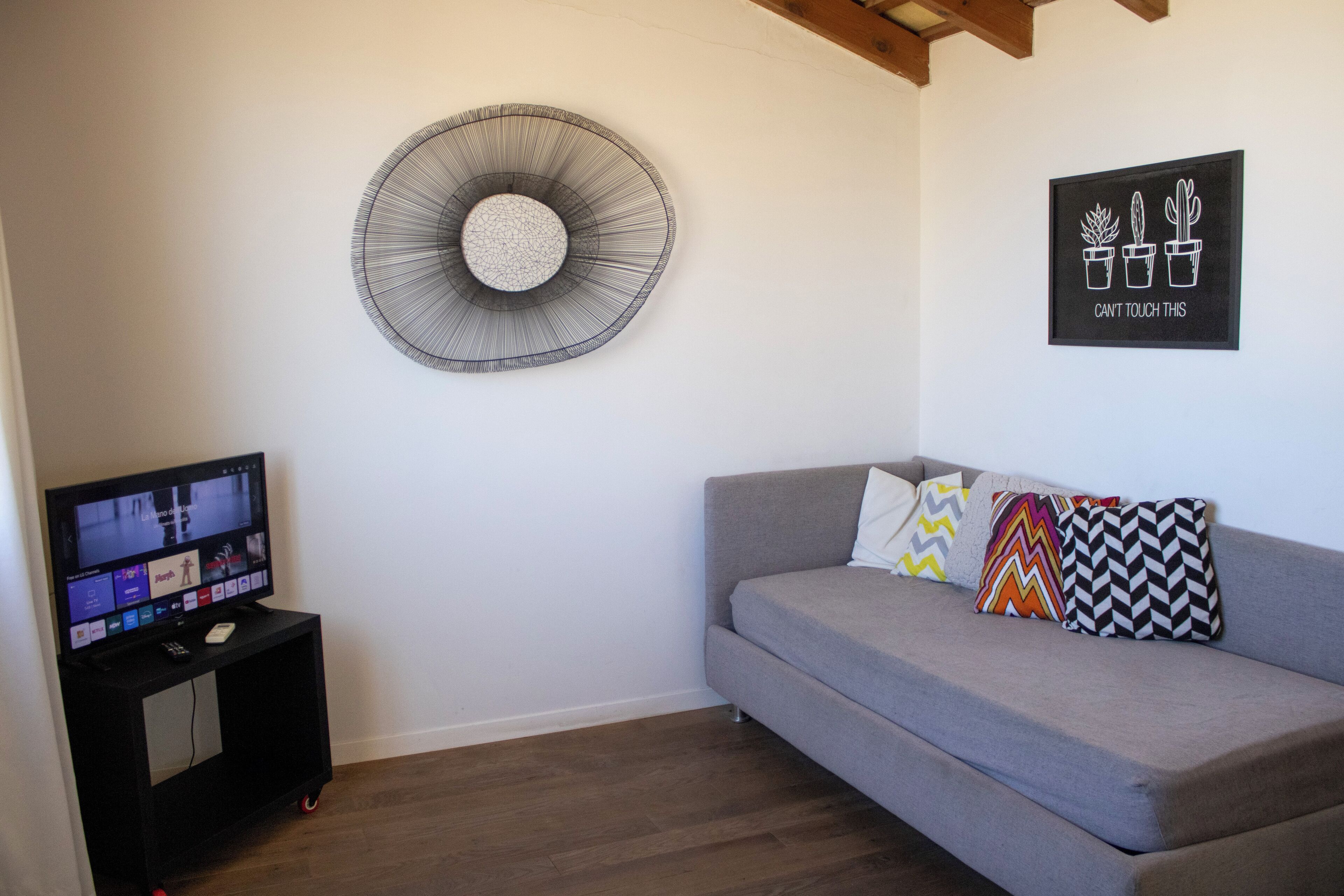 A living room with a grey sofa, colorful pillows, a wall-mounted fan, a flat-screen TV on a black stand, and a 'can't touch this' sign with potted plants.