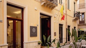 A building facade with a sign reading "BARDIA NOVA," outdoor seating with potted plants, and a flagpole with multiple flags. - BADIA NUOVA Apart Hotel (Trapani)