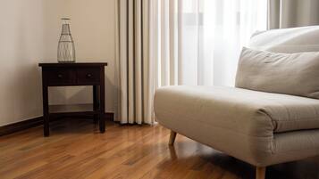 A room with a wooden floor, a beige sofa, a small wooden side table with a glass vase, and sheer white curtains.