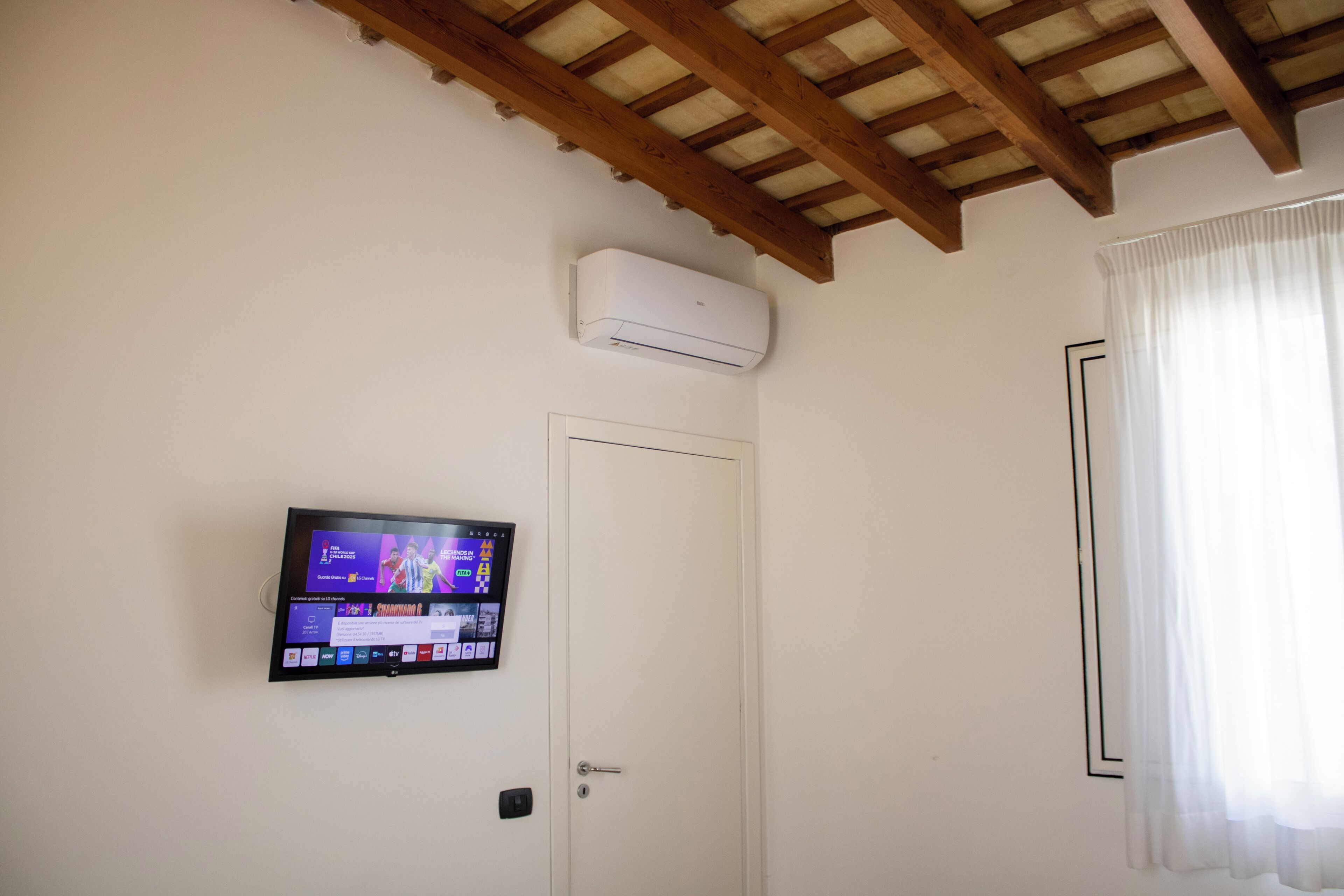 A room with a white door, a mounted air conditioner, and a flat-screen TV displaying a sports channel.