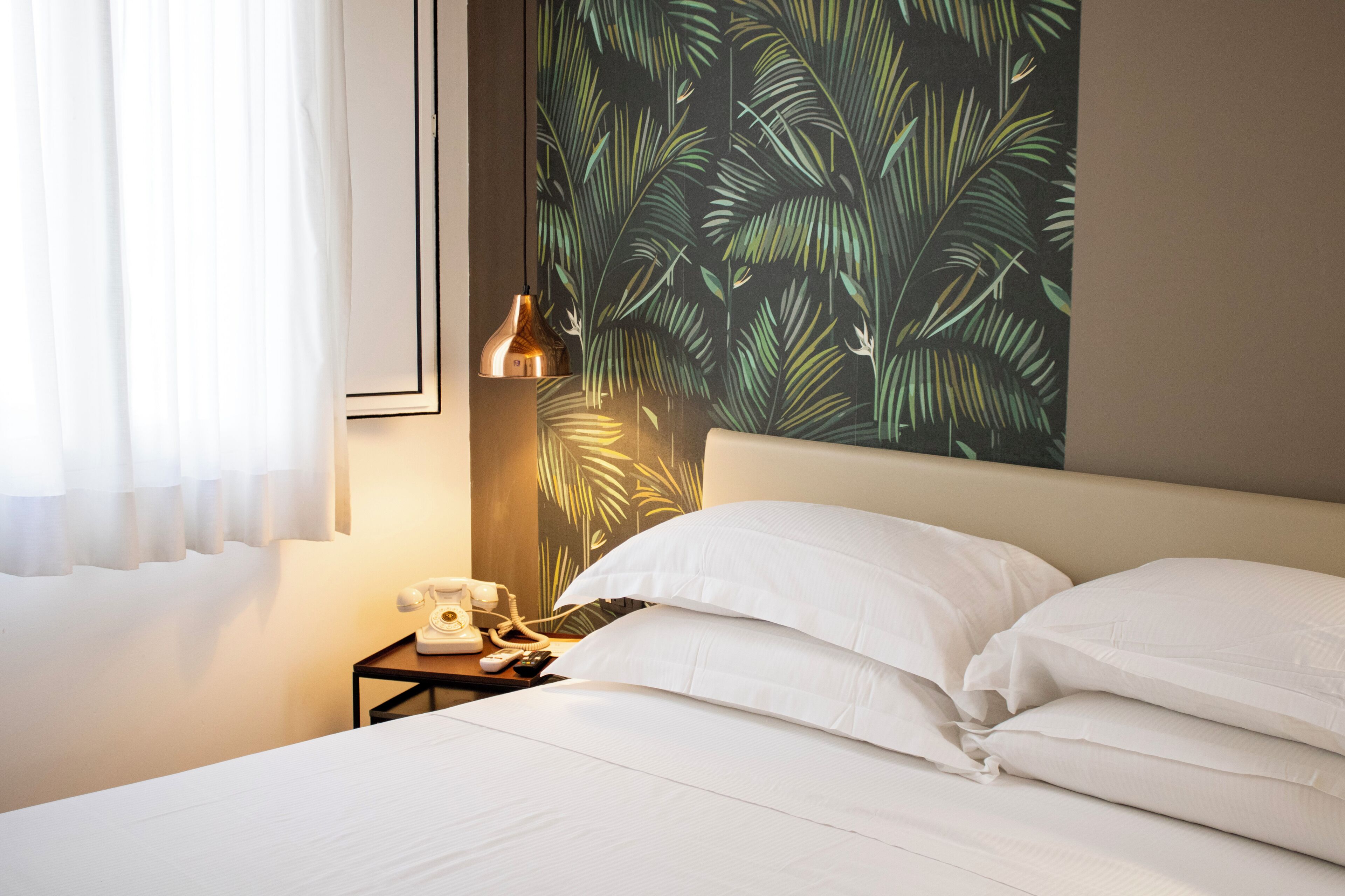 Une chambre d’hôtel avec un lit, des oreillers blancs, une table de chevet avec un téléphone et une télécommande, et un mur orné d’un motif de feuilles tropicales.