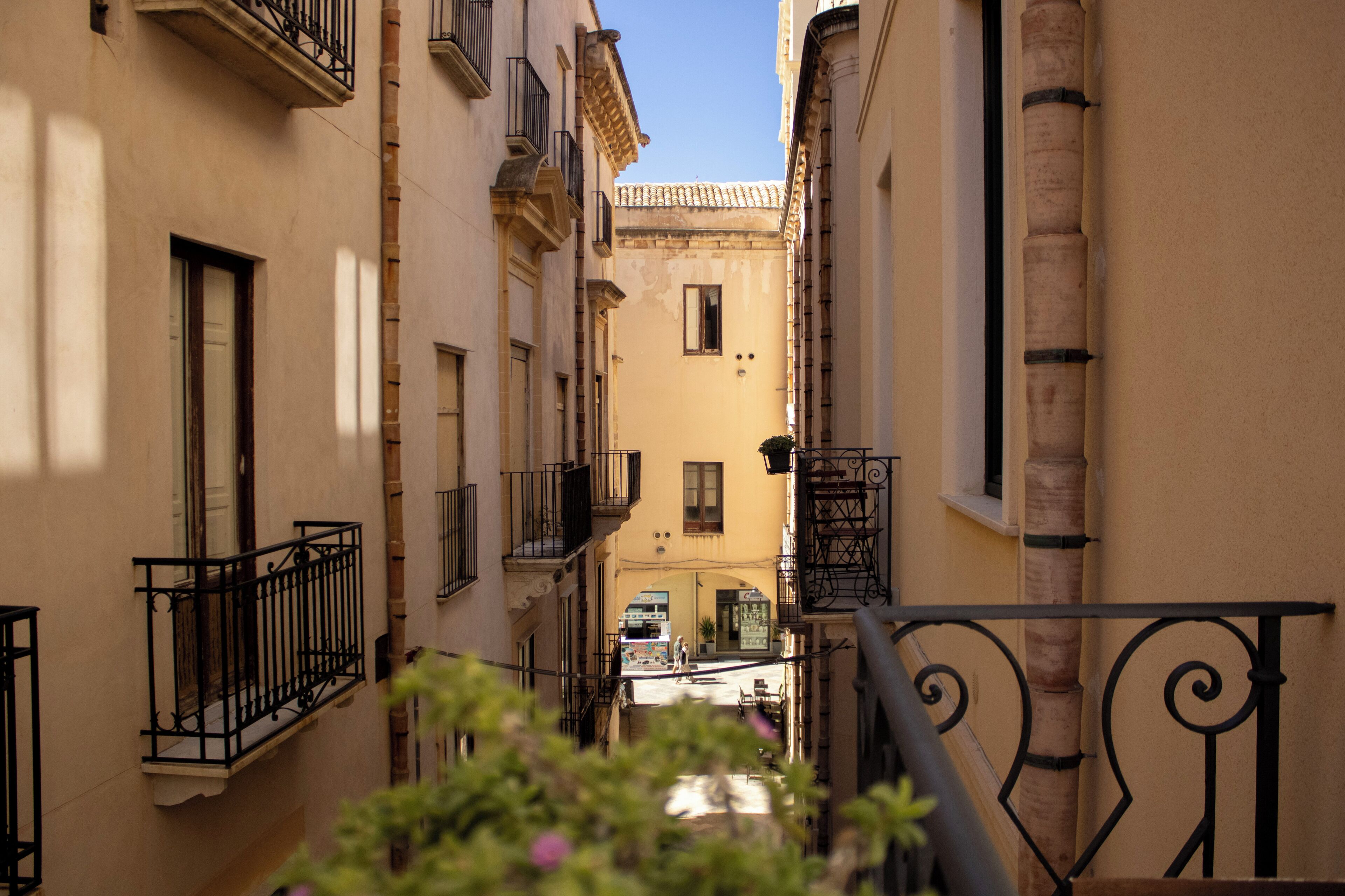A narrow street with buildings on either side, featuring balconies and a clear sky.