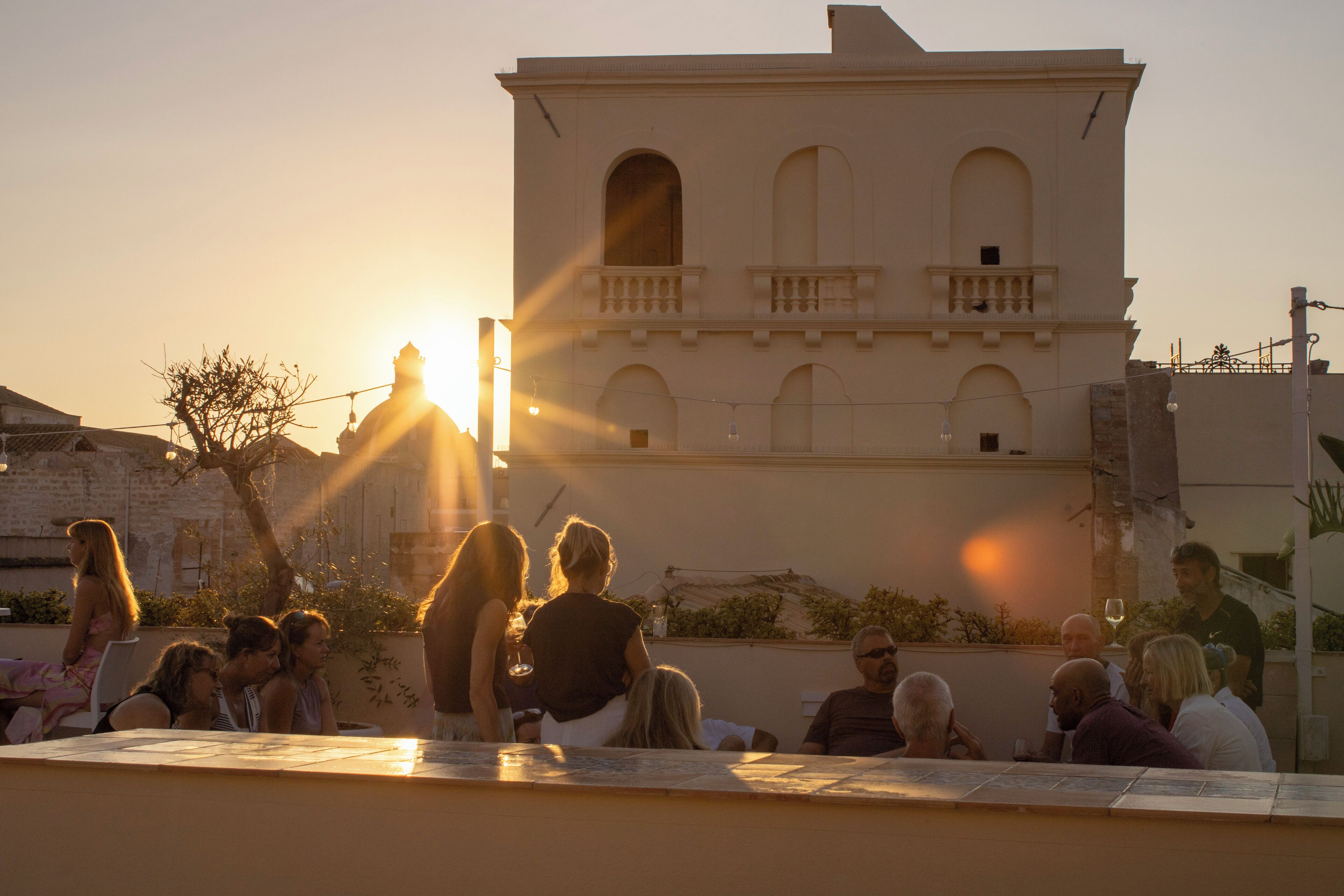 Un groupe de personnes est assis à l’extérieur, au coucher du soleil, devant un bâtiment doté d’arcatures et d’un balcon en arrière-plan.