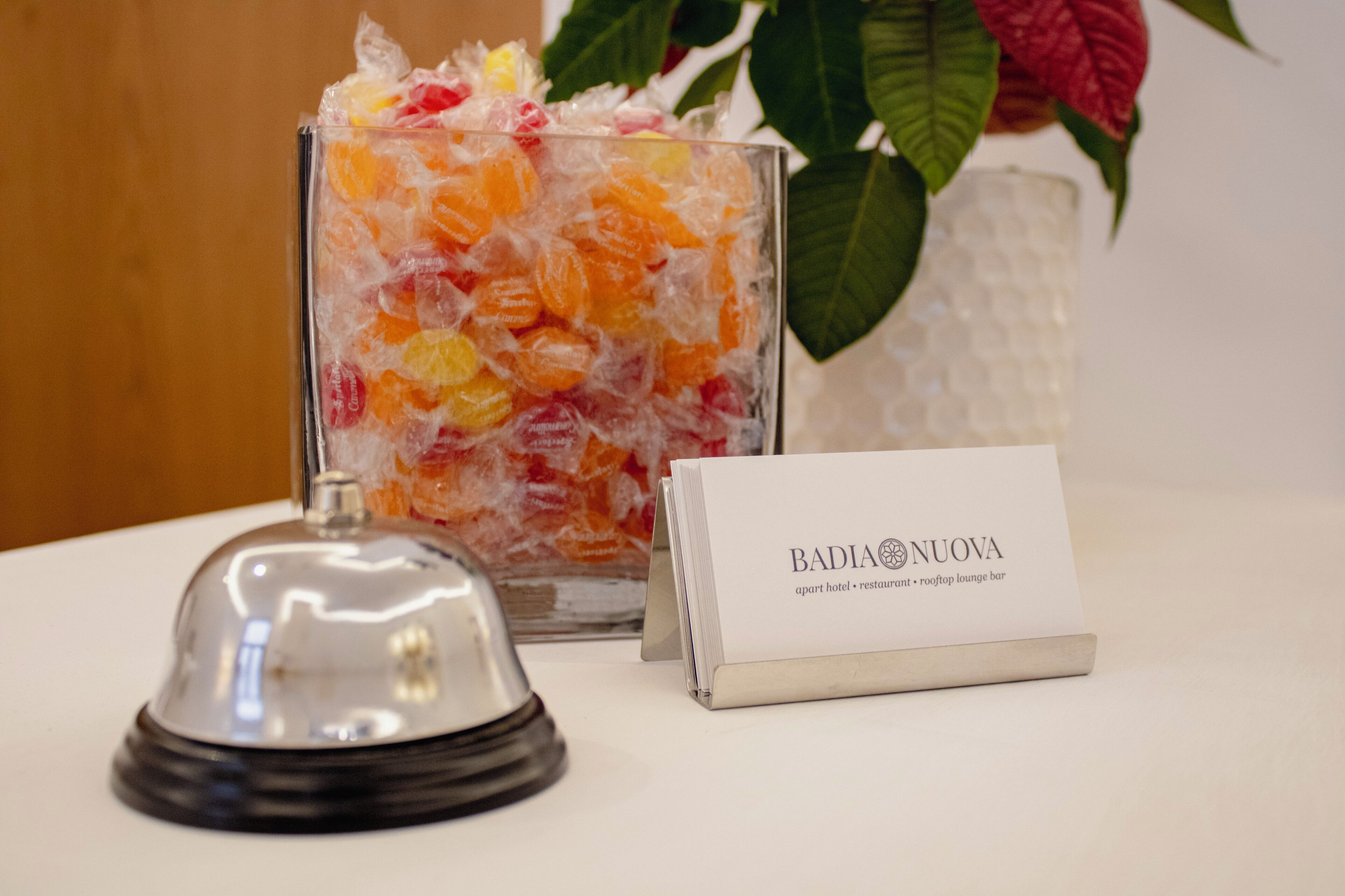 A glass container filled with colorful candies, a metal bell, and a business card for 'BADIA NUOVA' on a white surface.