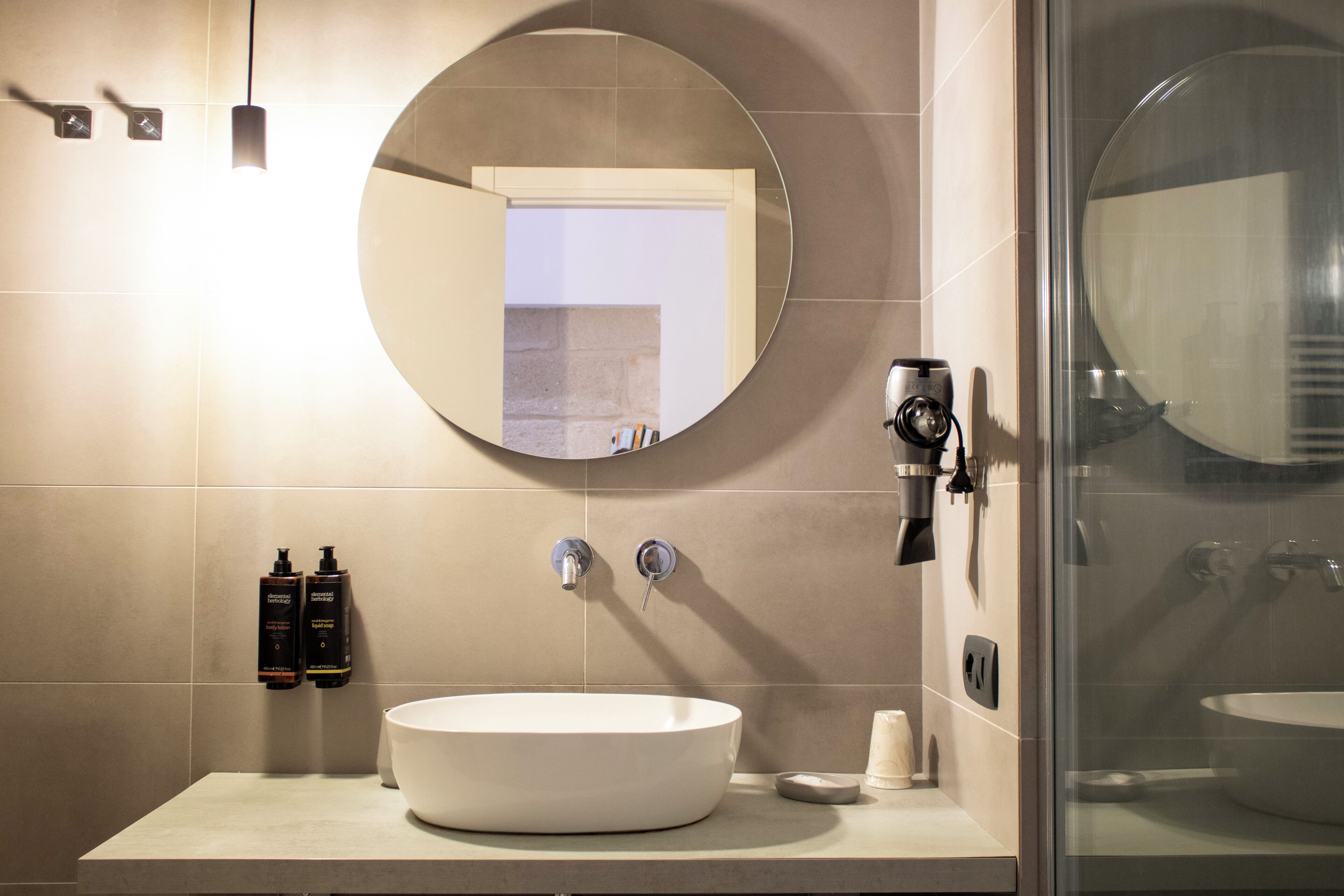 A modern bathroom with a large round mirror, a white sink, and two bottles of shampoo.