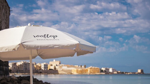 A beach umbrella with the text "rooftop" and "BADIA NUVONA" on it, set against a coastal backdrop with buildings and rocky shoreline. - BADIA NUOVA Apart Hotel (Trapani)