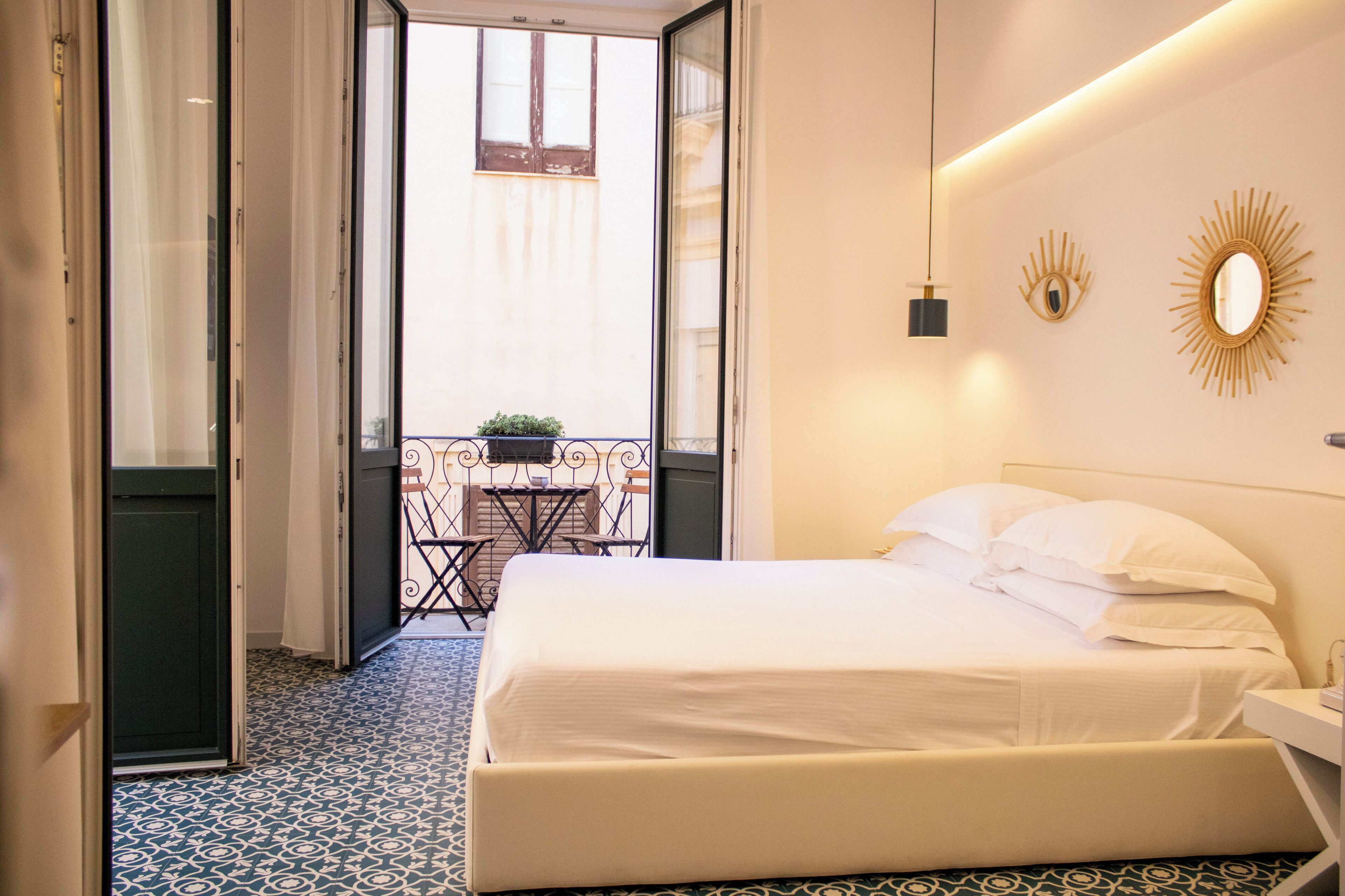 A hotel room with a bed, a small table with chairs, a balcony with a plant, and decorative wall mirrors.