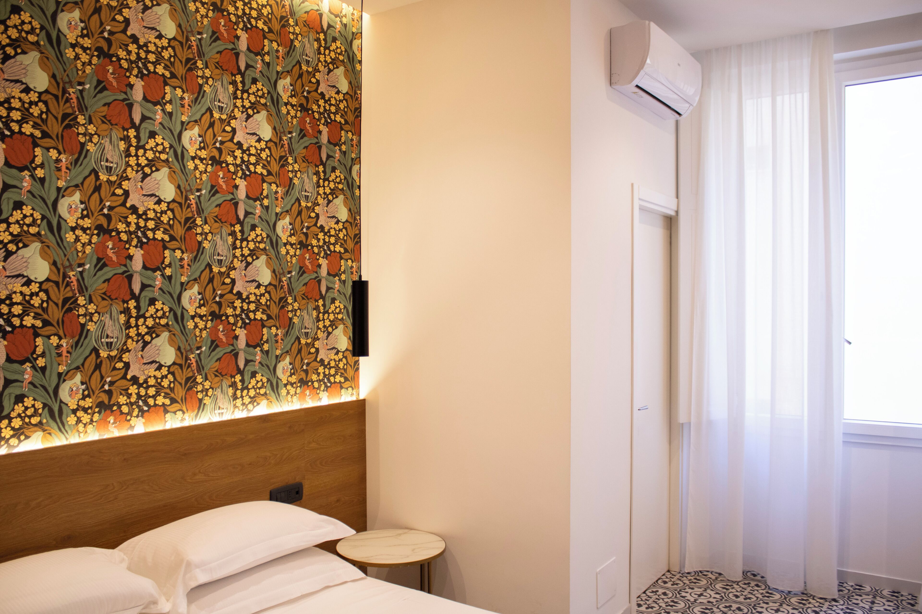 Ett hotellrum med en sänggavel med blommönster, ett litet runt bord, en säng med vita sängkläder och ett fönster med tunna, genomskinliga gardiner.