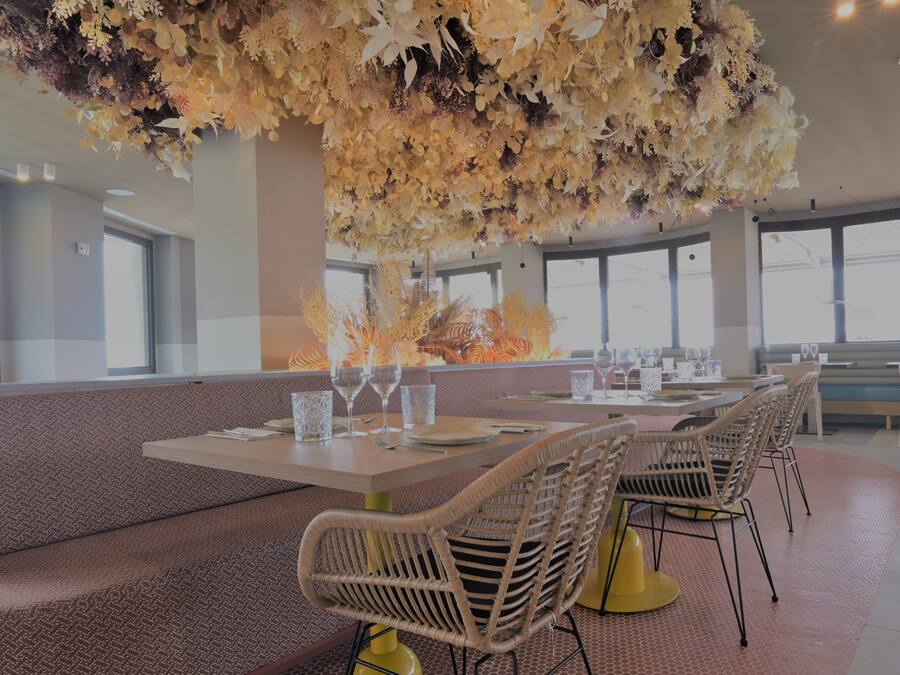 Un restaurante con techos altos, ventanas amplias y un arreglo floral central. Las mesas están dispuestas con sillas y copas de vino.
