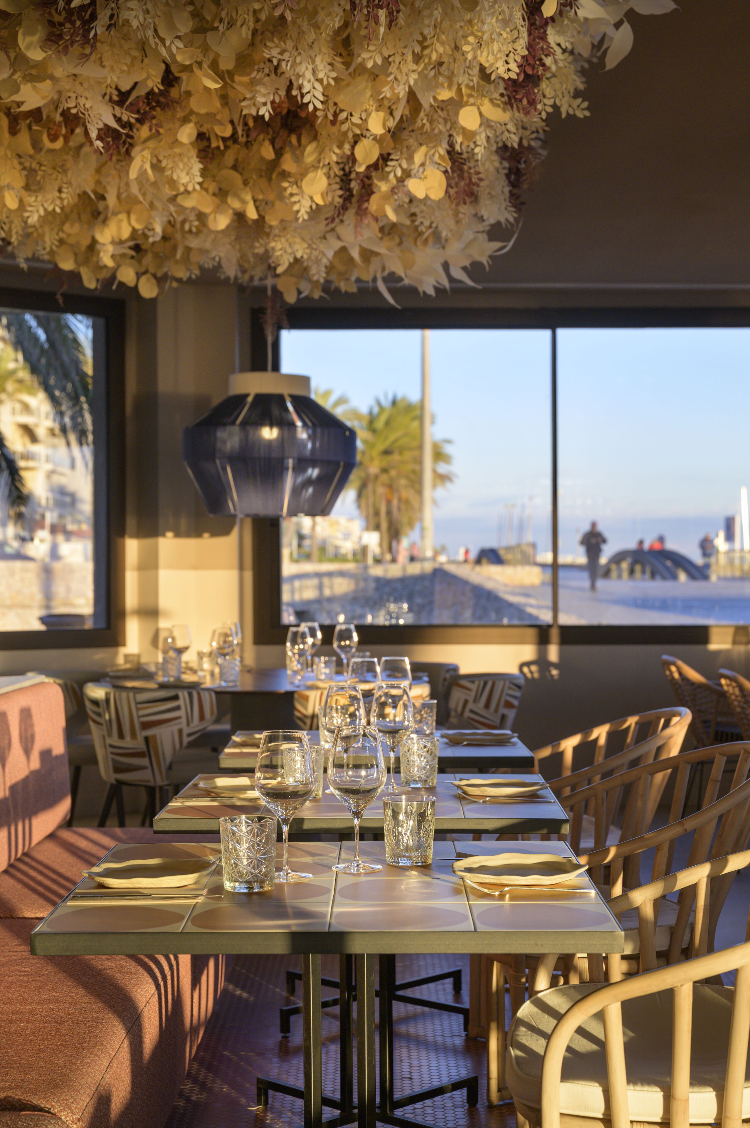 Un restaurante con una mesa lista para comer, un arreglo floral colgante y vistas a la playa a través de amplios ventanales.