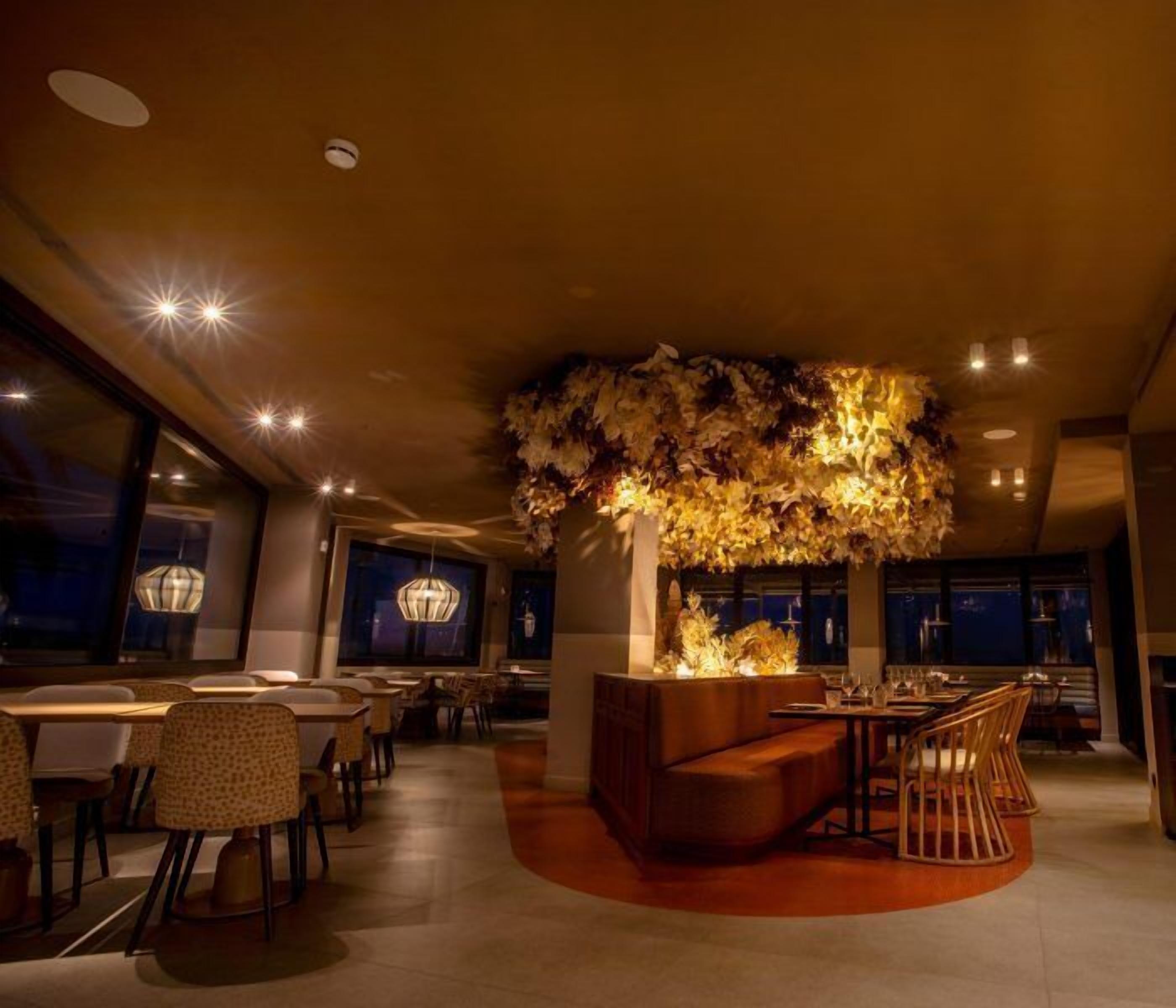 Un interior de restaurante moderno con una zona de estar central, mesas, sillas y un elemento decorativo en el techo.