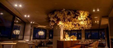 A modern restaurant interior with a central seating area, tables, chairs, and a decorative ceiling feature.