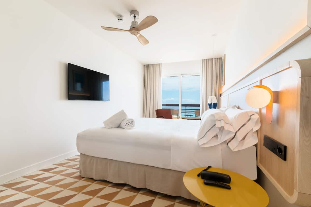 A hotel room with a bed, a ceiling fan, a TV mounted on the wall, a bedside table with a lamp, and a view of the sea through large windows.