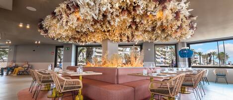 A restaurant with a central fire feature, cushioned seating, and a view of palm trees.
