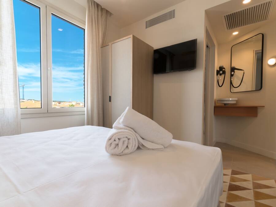 Habitación de hotel con cama, televisión, espejo y baño con lavamanos y toallero.