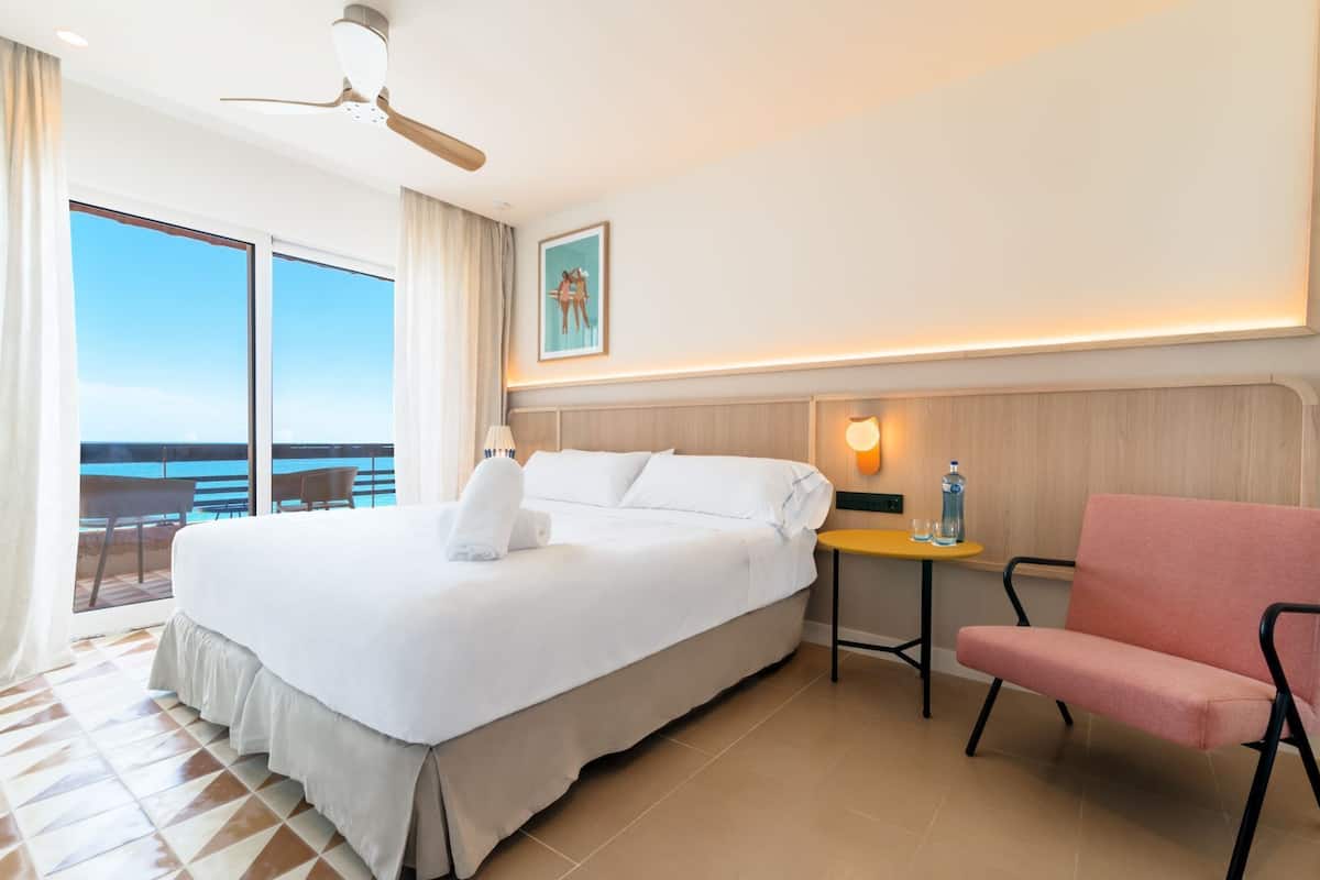 A hotel room with a large bed, a small table with a bottle and glasses, a chair, a ceiling fan, and a large window with a view of the ocean.