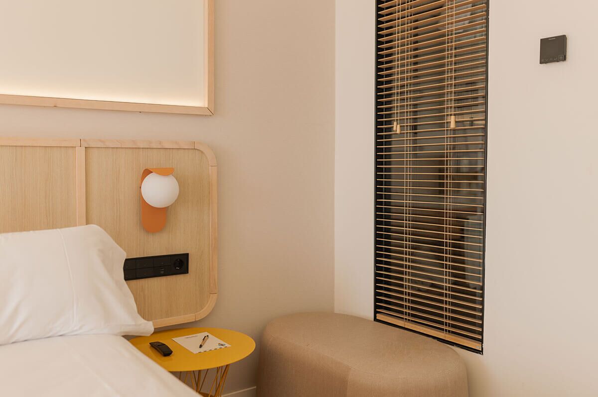 A hotel room with a wooden headboard, a bedside table with a lamp, a small round table with a phone and papers, and a window with blinds.