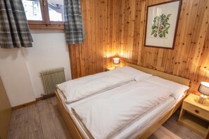 Cama doble con sábanas blancas, cabecera de madera, mesita de noche con lámpara e ilustración botánica enmarcada en la pared.