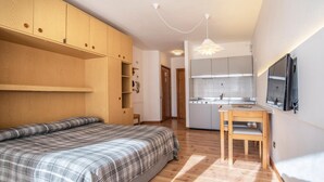 Une chambre compacte comprenant un lit, une armoire en bois, un coin cuisine et un espace repas.