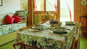 Une cabane chaleureuse avec un intérieur en bois, une table à manger dressée avec des assiettes et des bols, un canapé recouvert d’un tissu à motifs floraux et un téléviseur posé sur un meuble en bois.