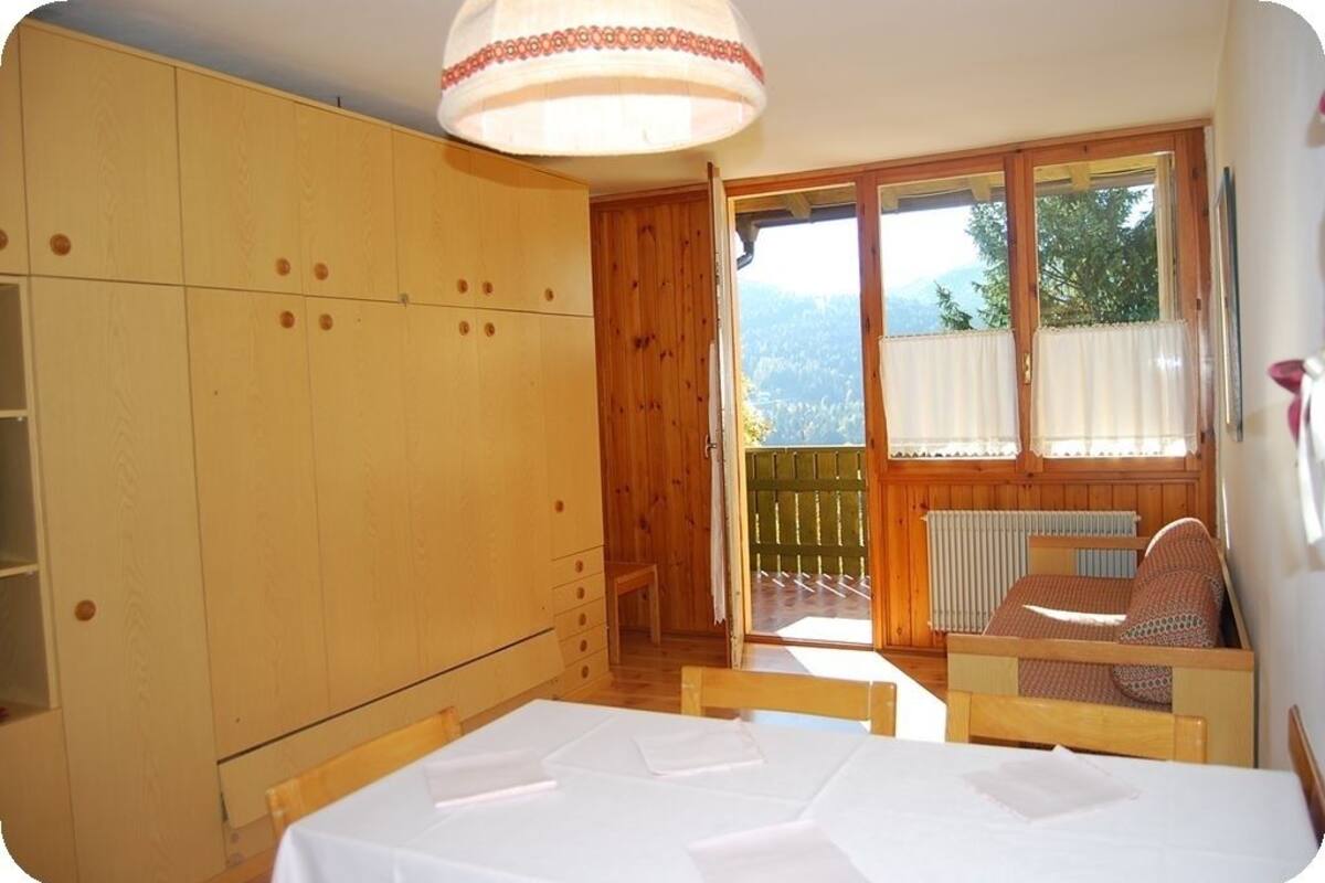 A room with a wooden wardrobe, a bed with a white mattress, a wooden table, and a window with a view of greenery.