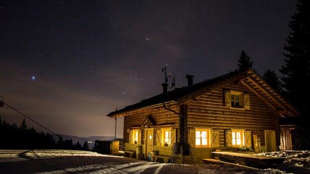 Une cabane en rondins avec un porche, des fenêtres illuminées et un ciel nocturne étoilé.