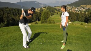 Un terrain de golf avec une chaîne de montagnes en arrière-plan, un golfeur en plein élan et un caddie qui se tient à proximité.