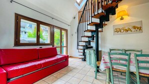 A room with a red sofa, a dining table with chairs, a staircase, and a window with a view of greenery. - Residence villa Frejus (Bardonecchia)