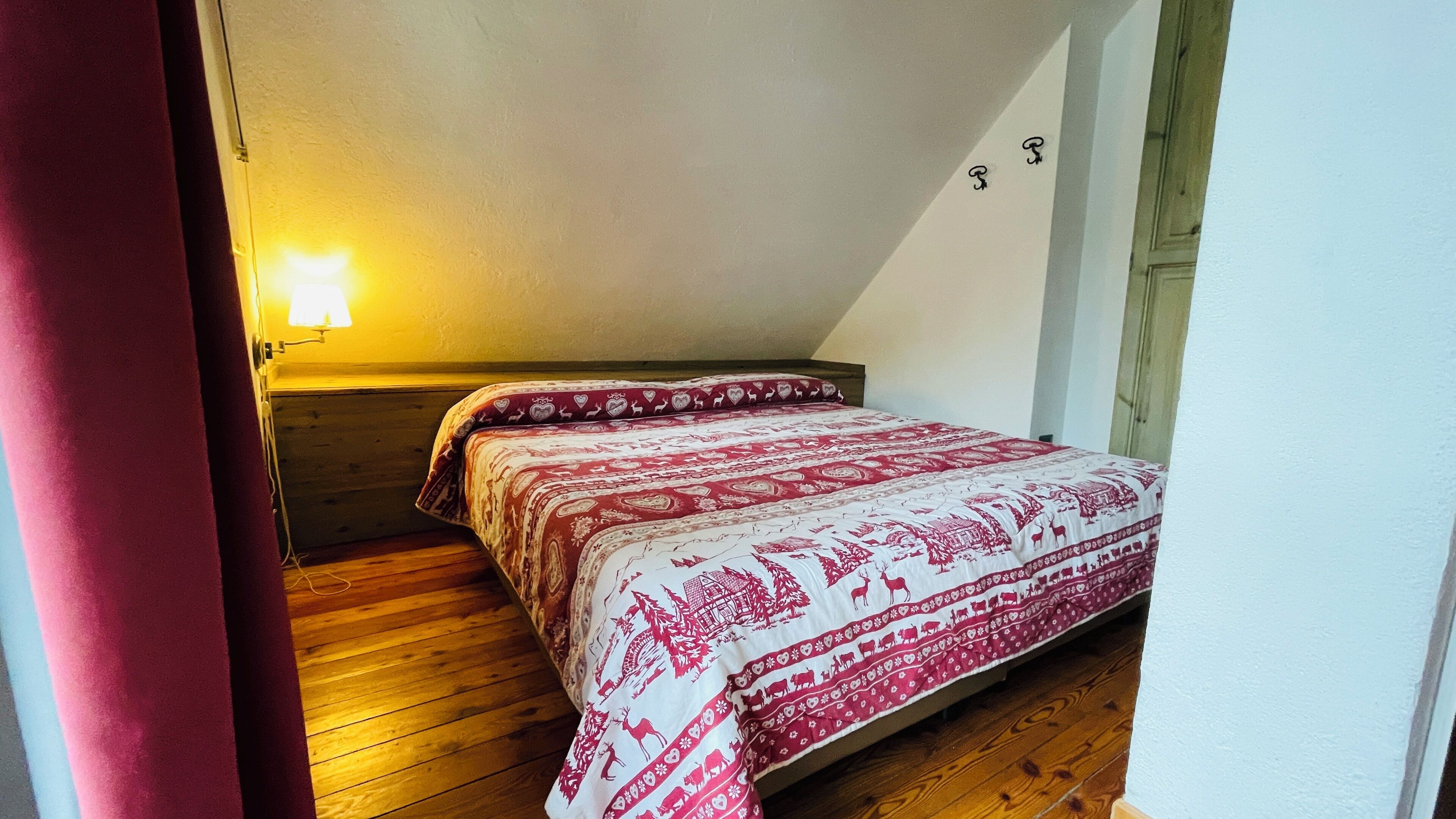 A small, cozy room with a wooden floor, a bed with a patterned bedspread, a nightstand, and a wall-mounted lamp.
