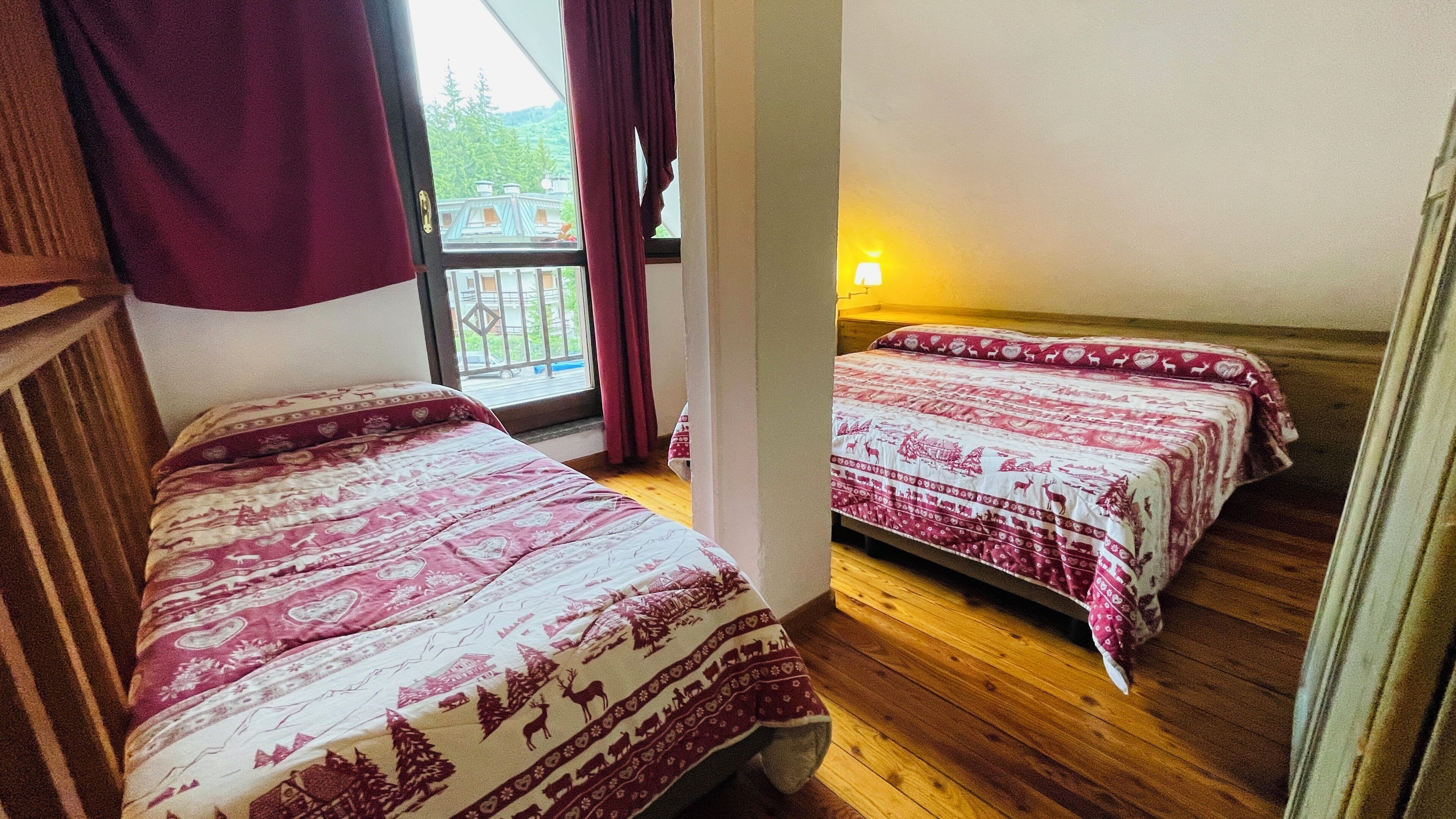 A room with two beds, wooden flooring, a window with a view of greenery, and a wall-mounted lamp.