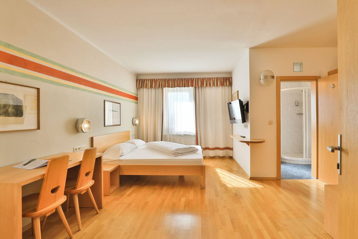 A hotel room with a wooden bed, a desk with chairs, a TV, and a shower area.