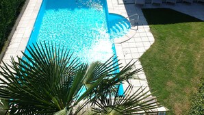 Una piscina rectangular con superficie de agua azul cristalina, rodeada por una terraza empavedada y una zona ajardinada.