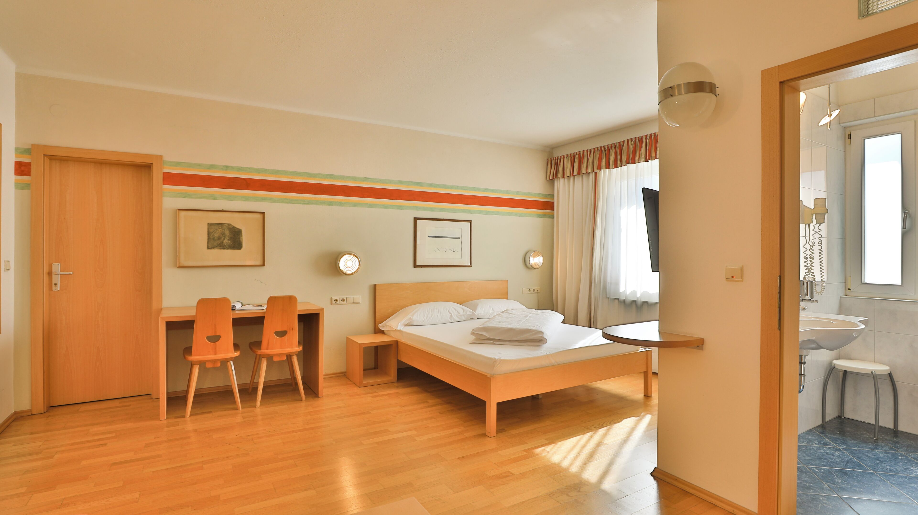 A hotel room with a wooden bed, a desk with two chairs, a bedside table, and a large window with curtains.
