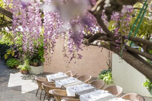 Un patio exterior con mesas y sillas de mimbre, rodeado de plantas en macetas y enredaderas florecientes.