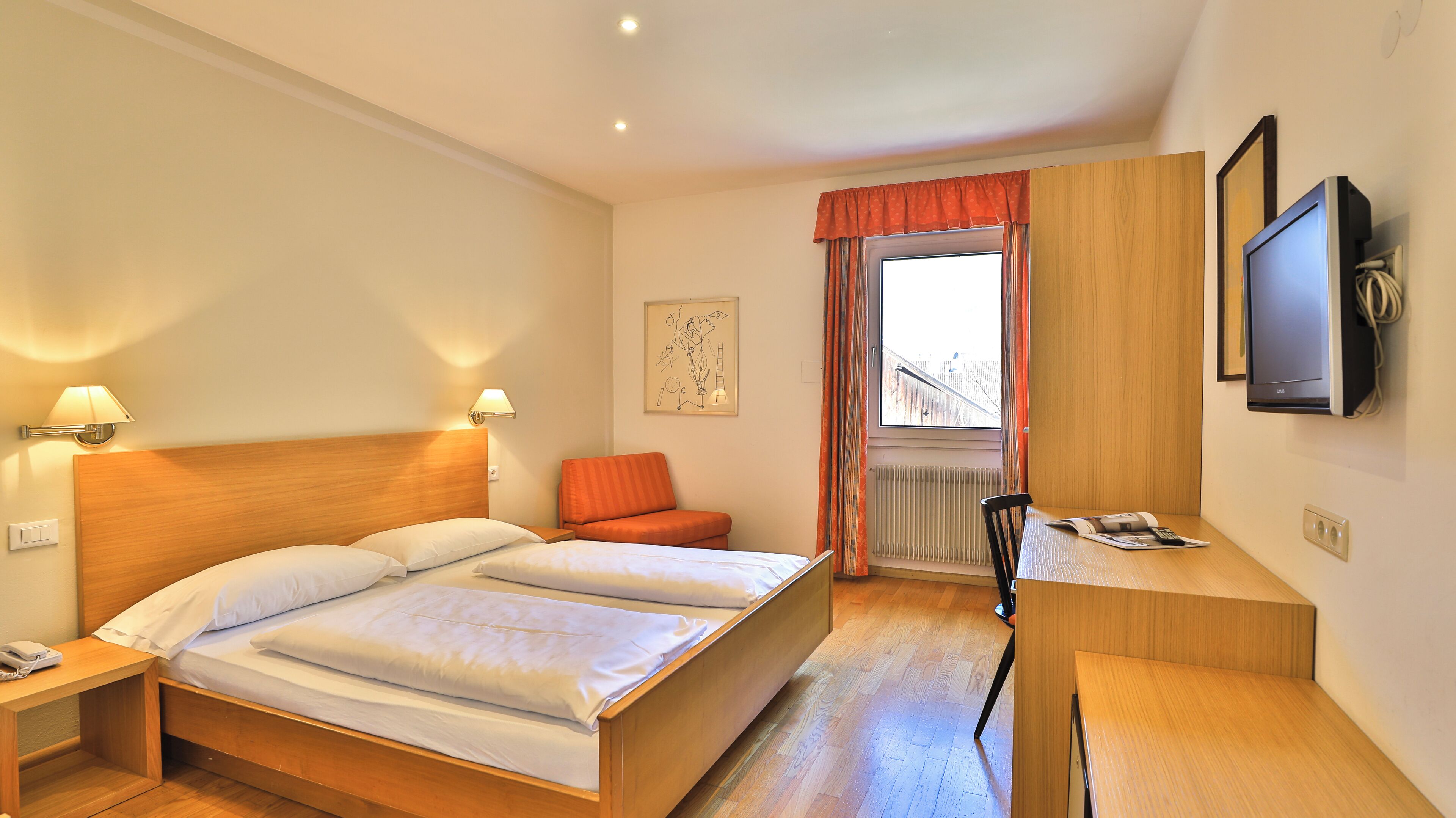 Een hotelkamer met een houten bed, twee nachtkastjes, een bureau, een stoel, een televisie en een raam met gordijnen.