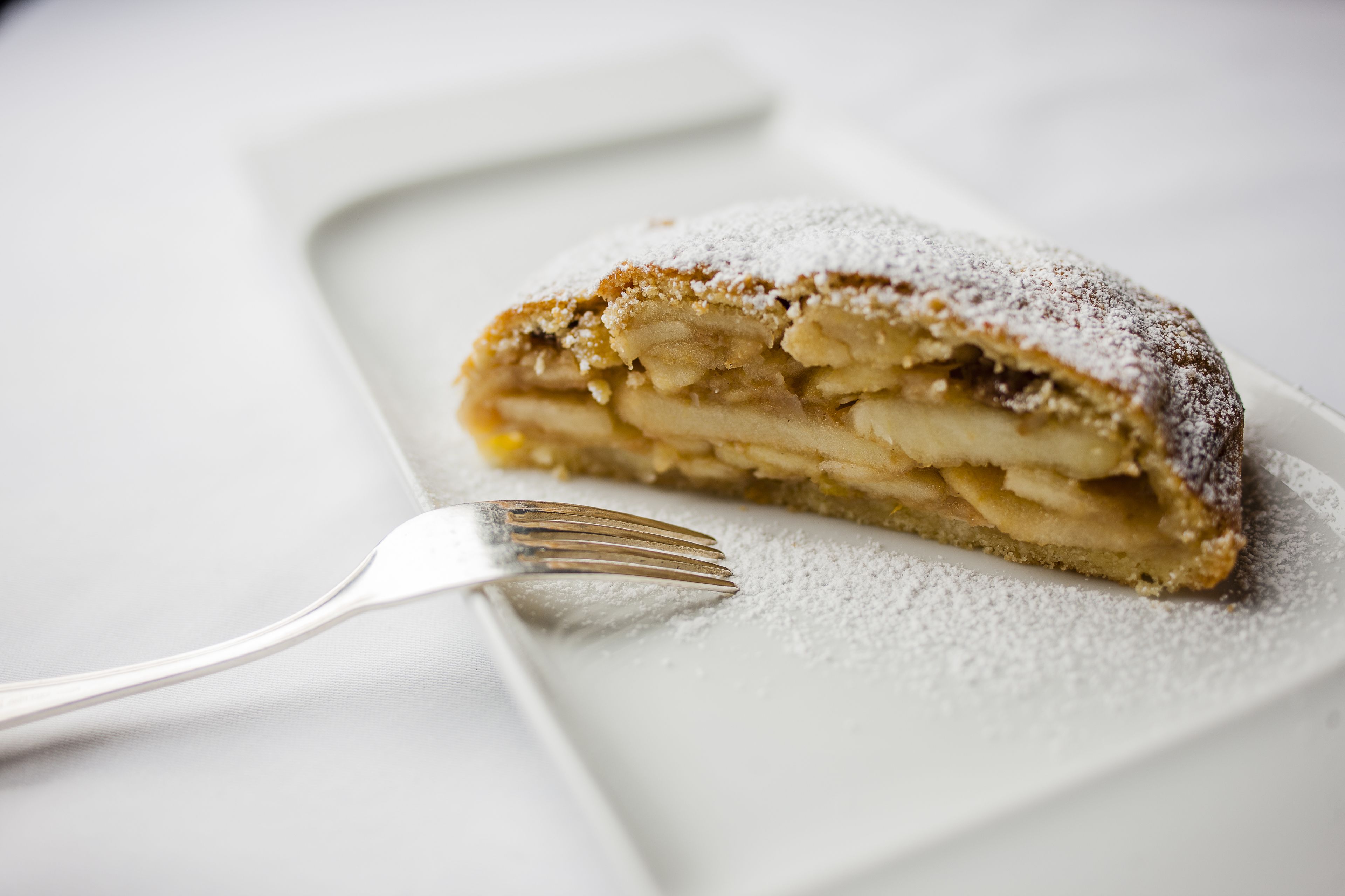 A slice of apple strudel on a white plate with powdered sugar, accompanied by a fork.