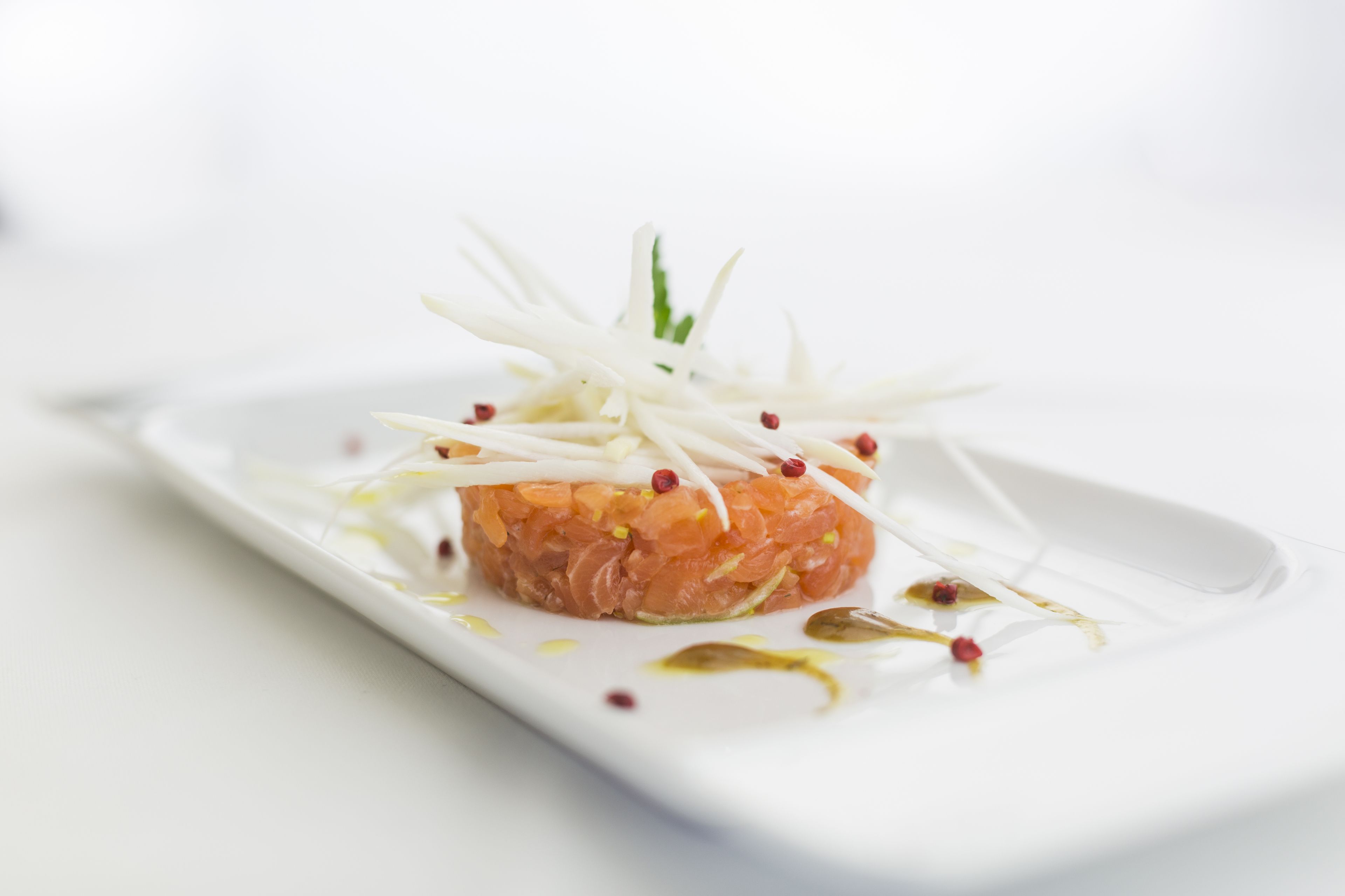 A white rectangular plate with a salmon tartare centerpiece, garnished with white shavings and red peppercorns, on a white background.