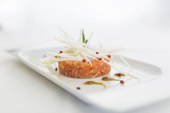 Plato blanco rectangular con un centro de tártara de salmón, adornado con virutas blancas y granos de pimienta roja, sobre fondo blanco.