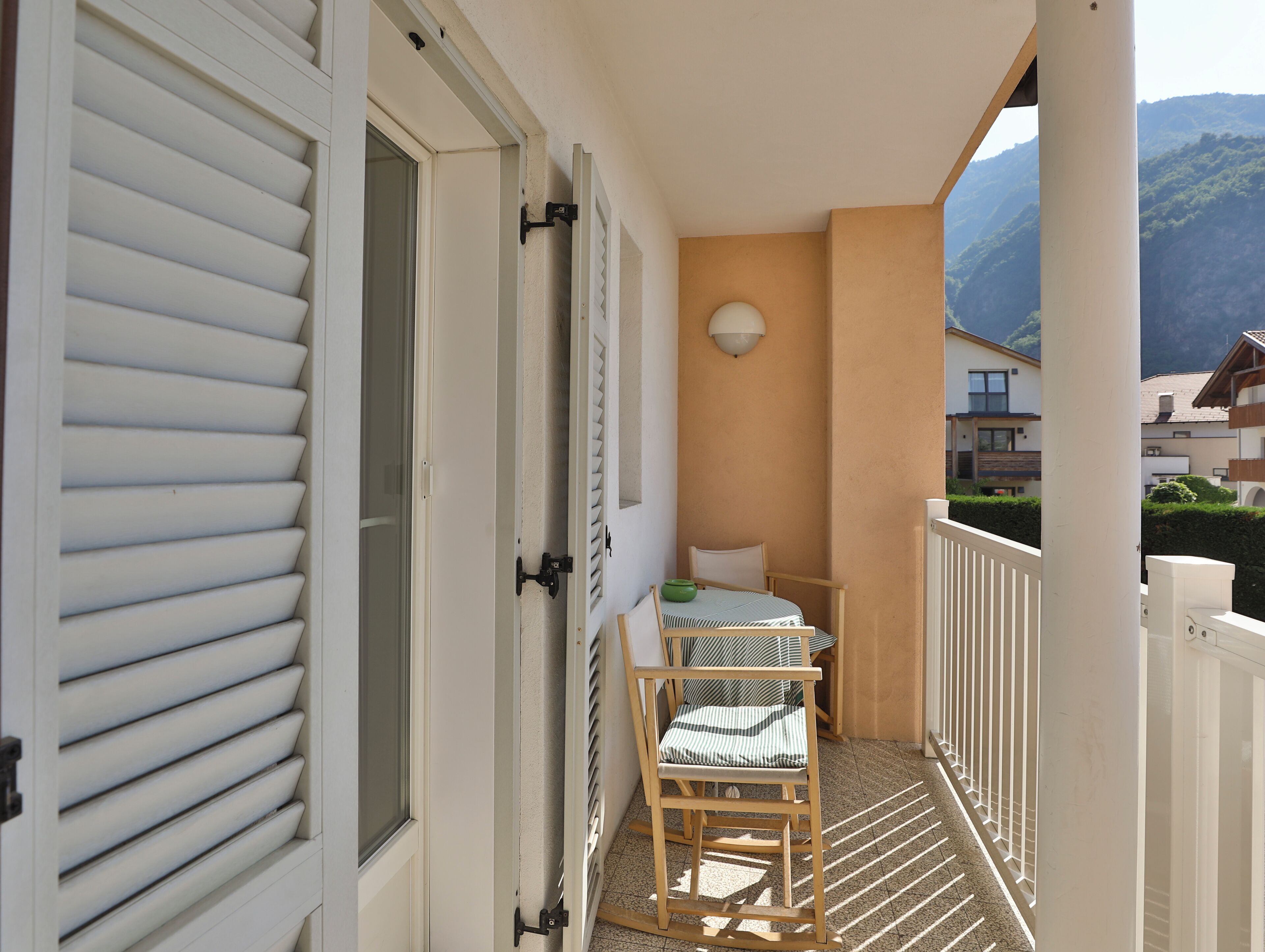 Ein Balkon mit einem Holzstuhl und einem kleinen Tisch, eine weiße Tür mit Fensterläden und Blick auf einen Berg.