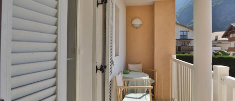 Ein Balkon mit einem Holzstuhl und einem kleinen Tisch, eine weiße Tür mit Fensterläden und Blick auf einen Berg.