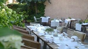 Un área de comedor al aire libre con mesas preparadas para una comida, rodeada de follaje y una pared color beige.