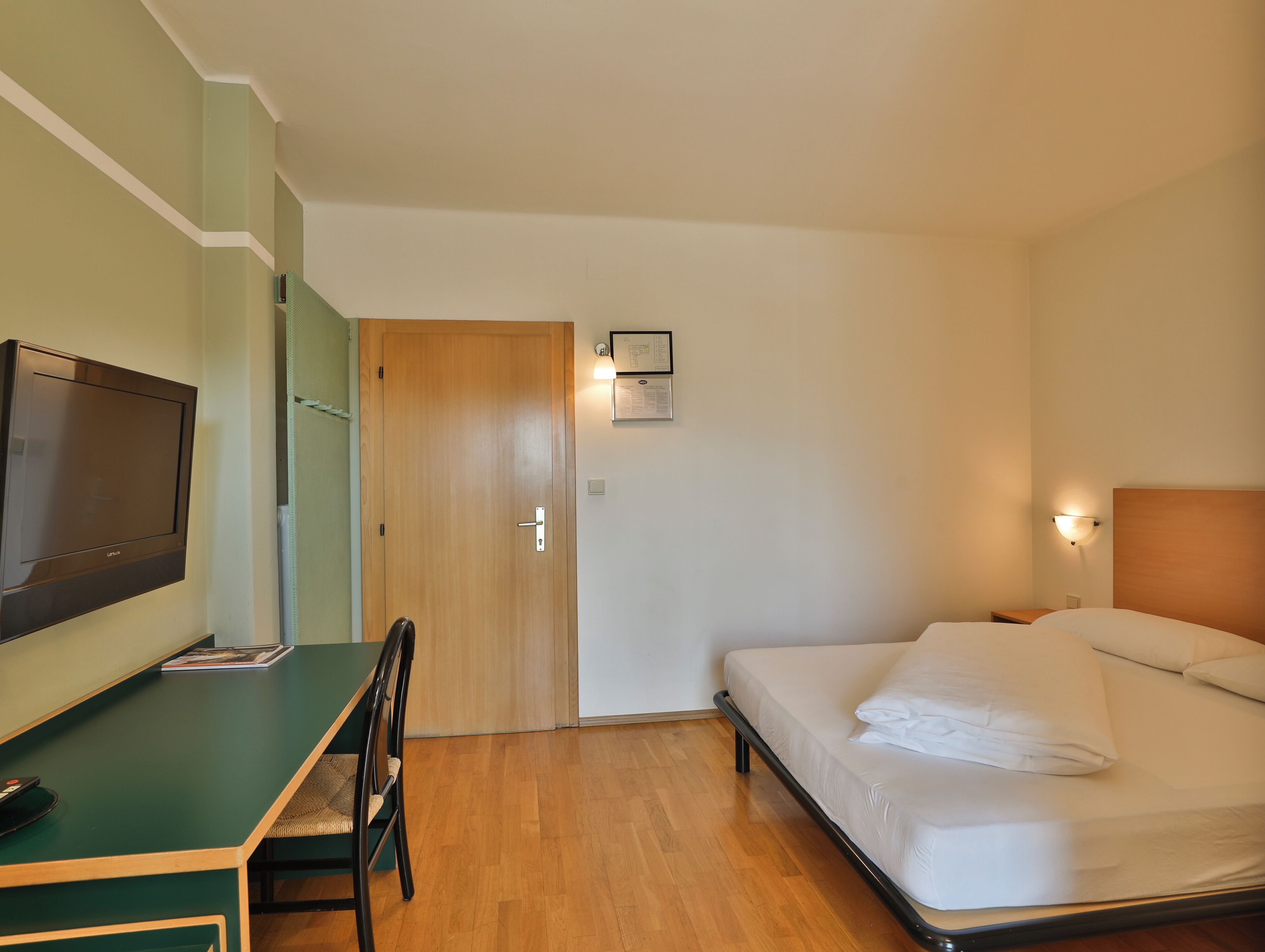 A hotel room with a bed, a desk with a chair, a television, and a wall-mounted light.