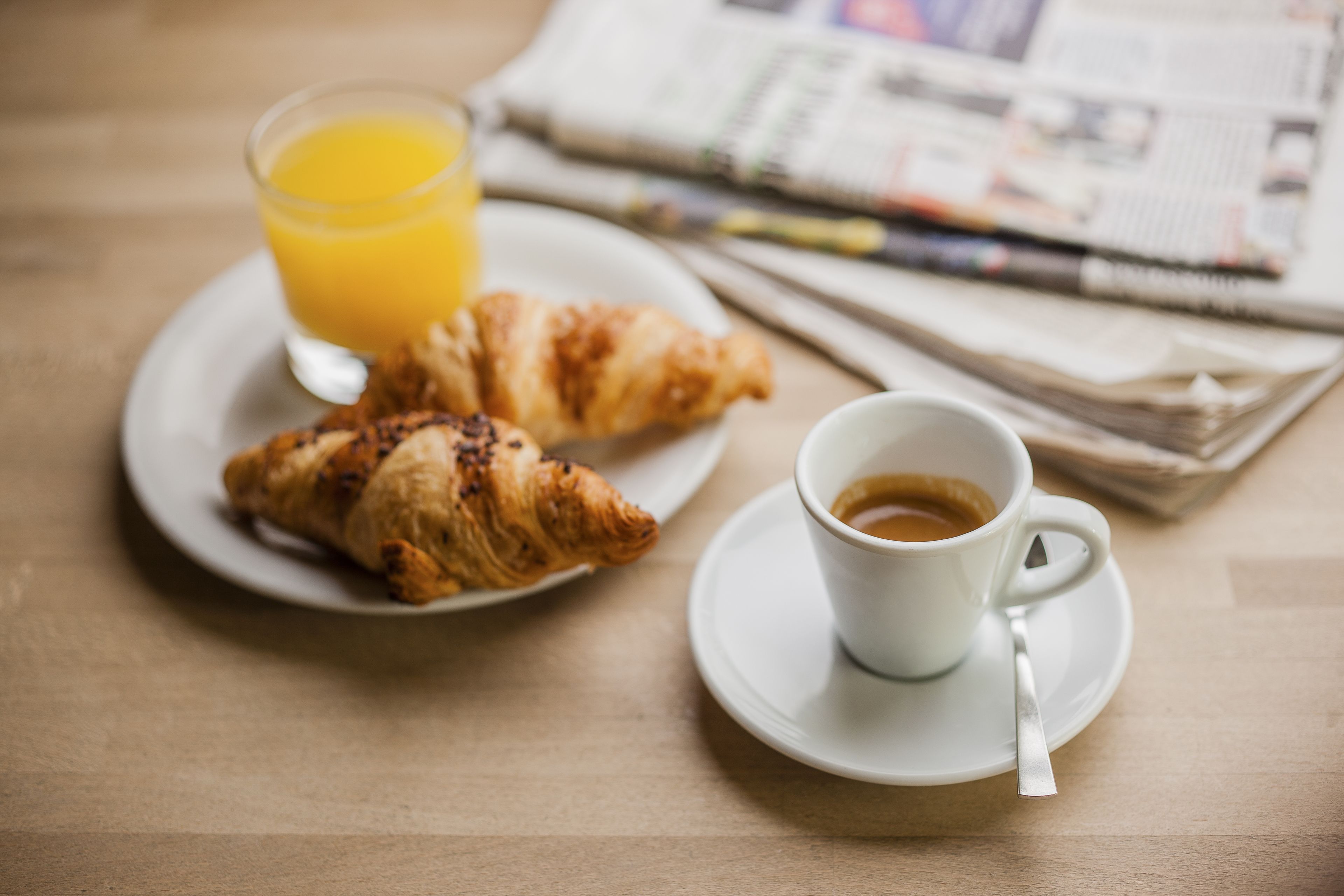 A breakfast setting with croissants, a cup of coffee, and a glass of orange juice on a table with newspapers.