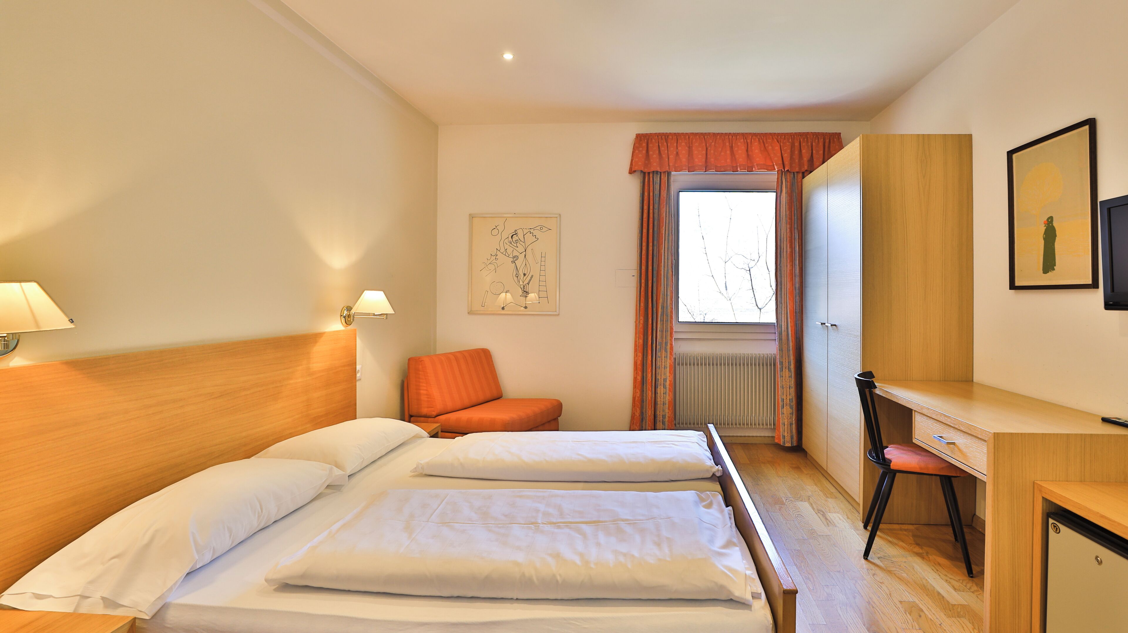 Ett hotellrum med en dubbelsäng, ett skrivbord, en orange stol, ett fönster med gardiner och en vägglampa.