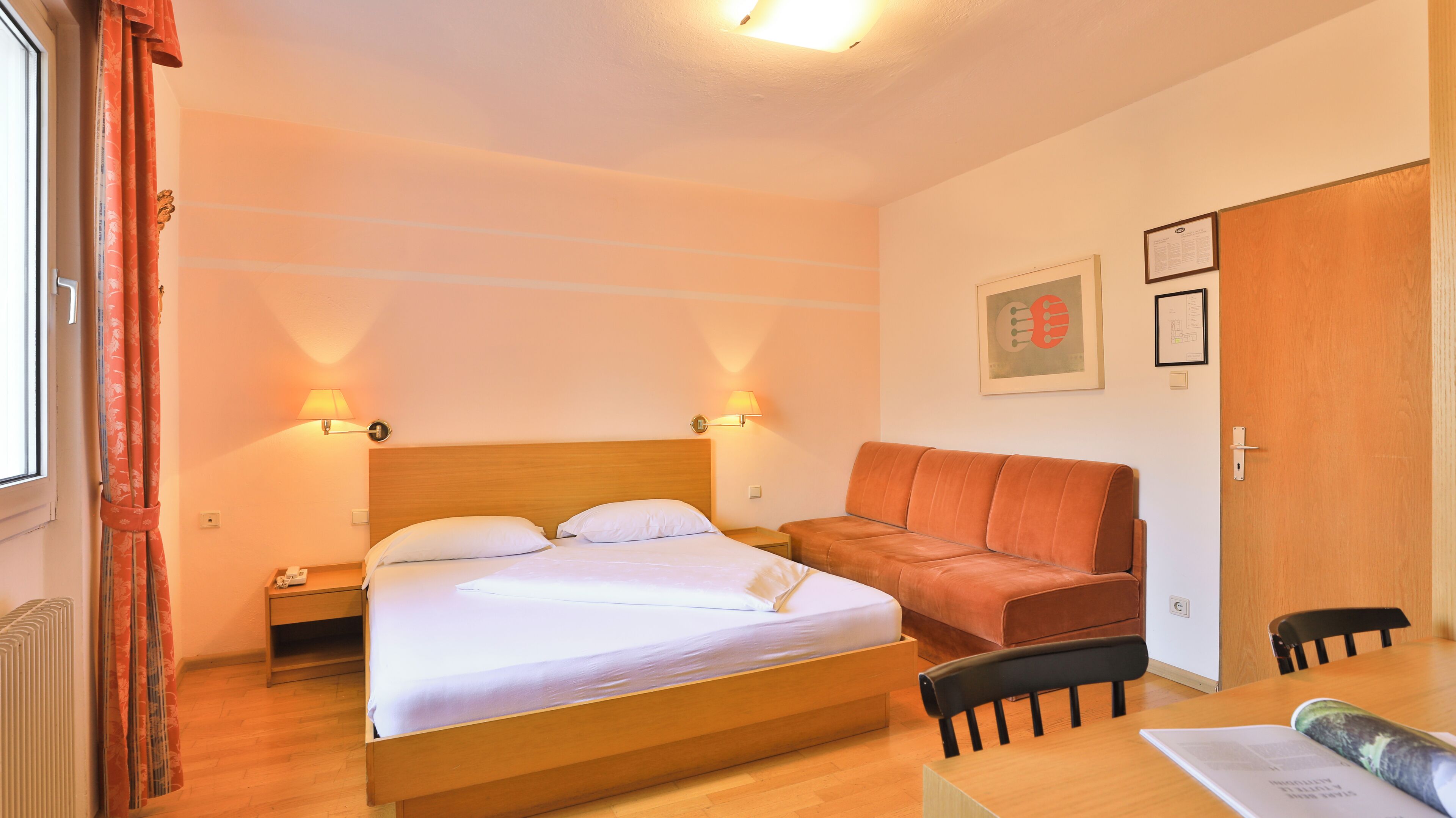 A hotel room with a wooden bed, two bedside lamps, a sofa, a table with chairs, and a door.