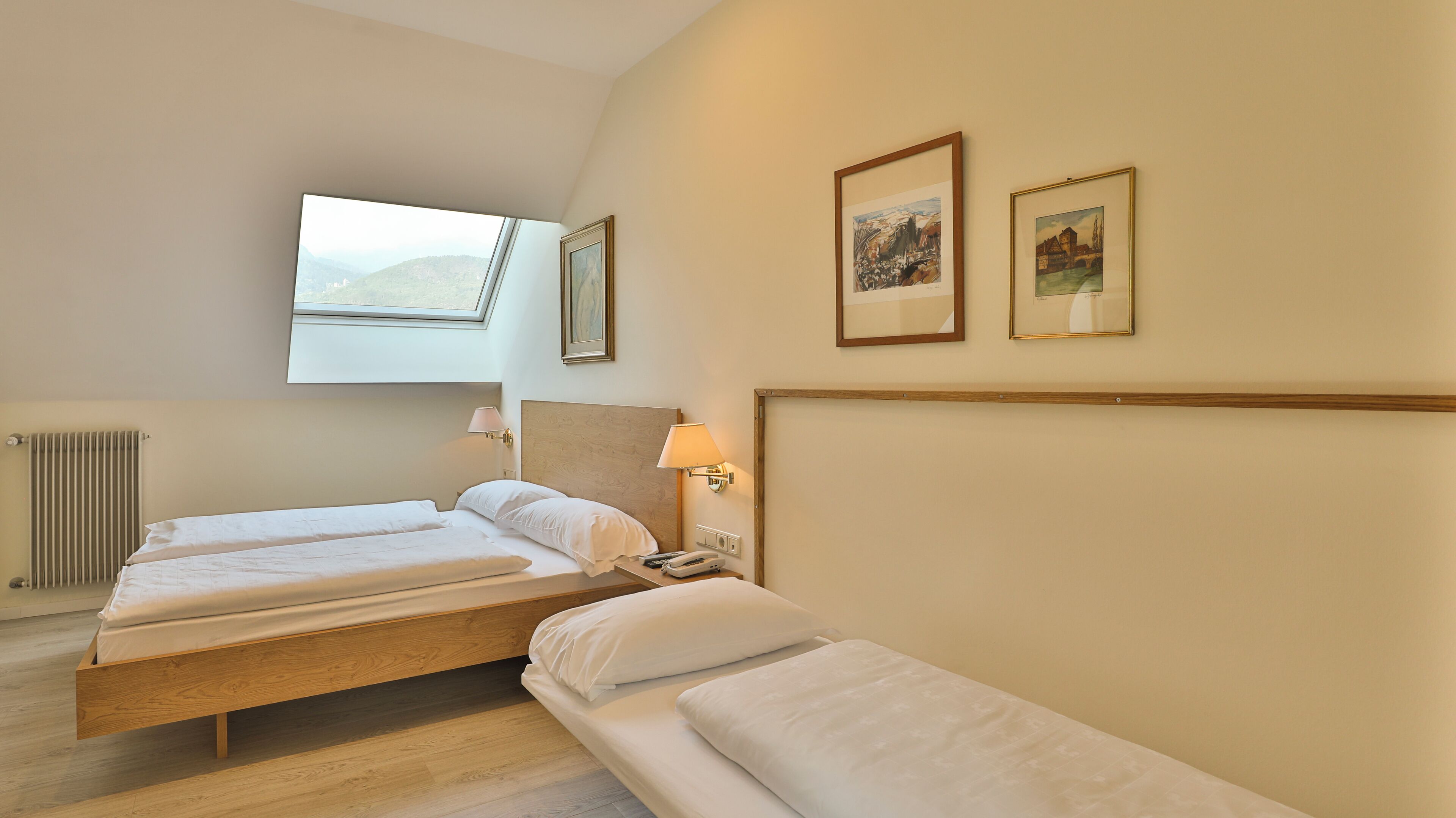 Ein Hotelzimmer mit zwei Betten, einem Oberlicht, einem Heizkörper und zwei gerahmten Bildern an der Wand.