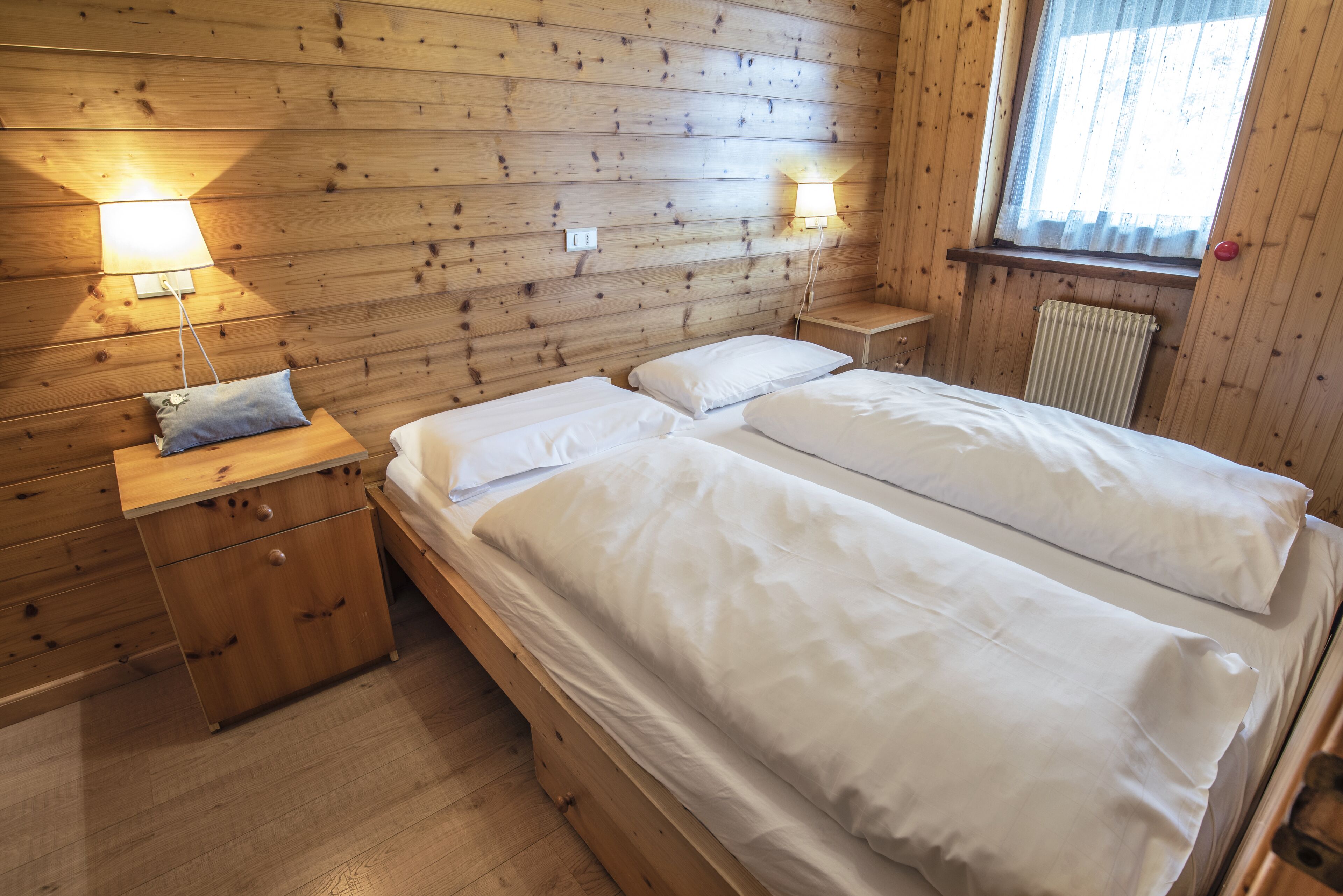 Una habitación de madera con dos camas, una mesita de noche, una lámpara y un ventanal con cortinas.
