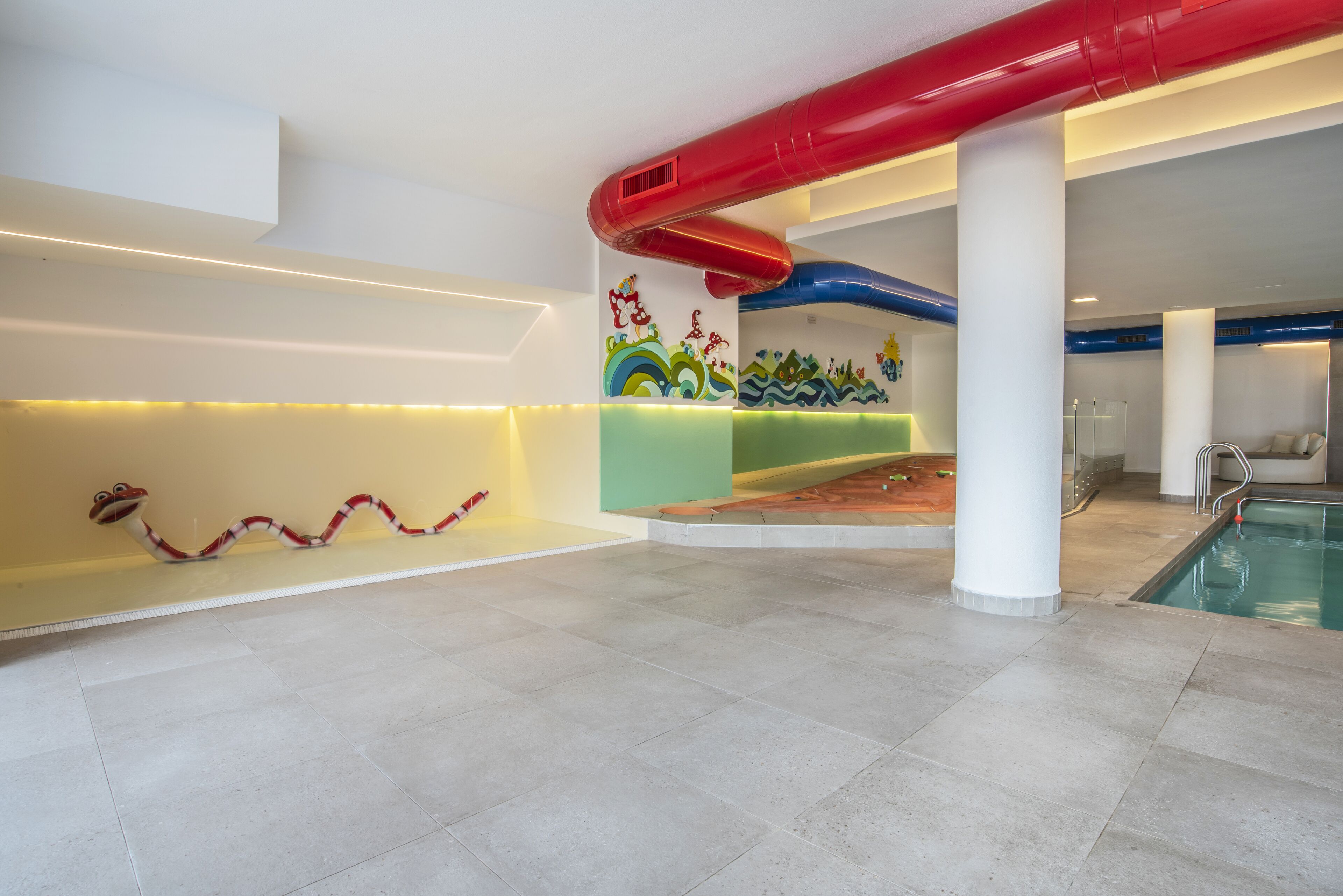 Un área de piscina moderna en interior con un tobogán rojo y azul, un mural colorido y una piscina.
