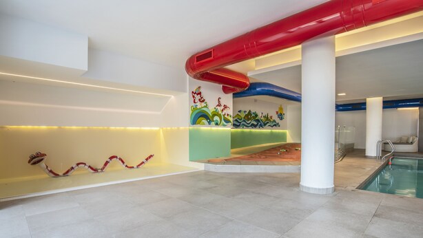 Un área de piscina moderna en interior con un tobogán rojo y azul, un mural colorido y una piscina.